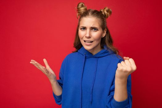 Photo of emotional agressive young beautiful blonde woman with sincere emotions wearing blue pullover isolated over red background with free space and showing fist