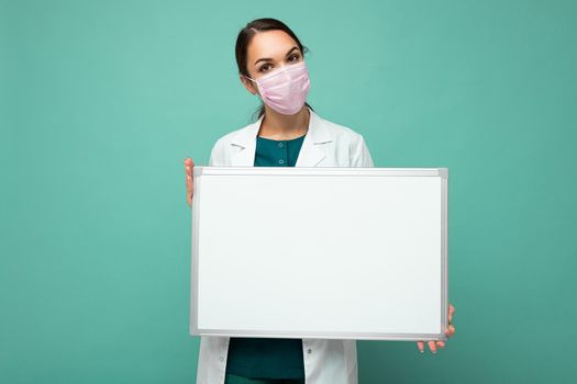 Woman doctor wearing a white medical coat and a mask holding blank board with copy space for text isolated on background. Coronavirus concept