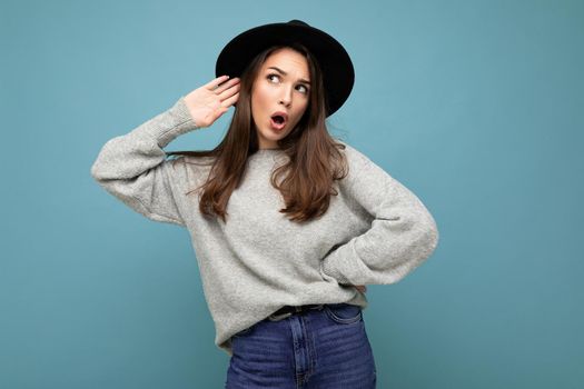 Portrait of young emotional beautiful brunette woman with sincere emotions wearing stylish grey pullover and black hat isolated over blue background with copy space and listening to gossip with shock expression