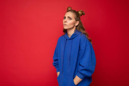 Portrait of sorrowful tired young attractive winsome blonde woman with two horns with sincere emotions wearing casual bright blue hoodie isolated on red background with free space