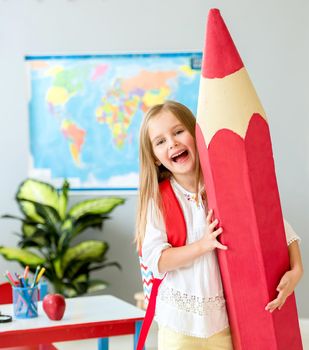 Little smiling blond girl holding huge red pencil in the school classroom