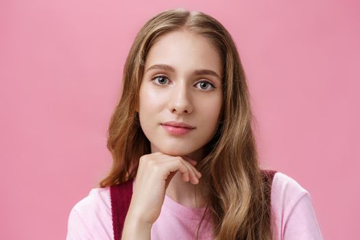 Close-up shot of charming glamourous young feminine girl with wavy natural hair and slight makeup holding hand on chin and smiling with dreamy friendly-looking sensual look over pink wall