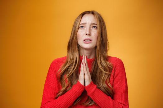 Close-up shot of anxious nervous and hopeless distressed cute redhead woman in red dress holding hands in pray looking up with supplication praying asking god help or mercy over orange wall