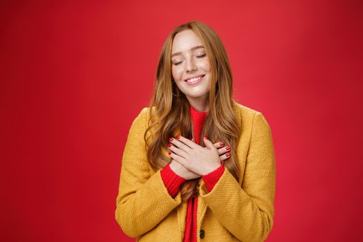 Girl feeling like heart warming as remembering sweet tender memories with closed eyes and silly broad smile holding hands on chest pressing to soul posing nostalgic and touched over red background