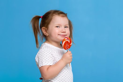 Beautiful little female child holding huge lollipop spiral candy smiling happy isolated on blue background.
