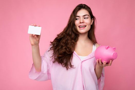 Portrait of beautiful positive cheerful cute smiling young brunette woman in stylish shirt isolated on pink background with copy space and holding pink pig moneybox and credit card for mockup