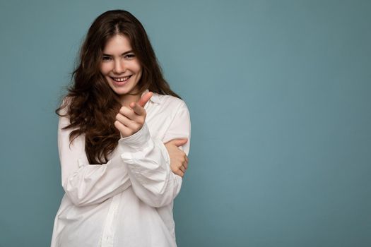 Portrait of young positive happy smiling cite nice curly brunette woman with sincere emotions wearing casual white shirt isolated on blue background with empty space and pointing at camera