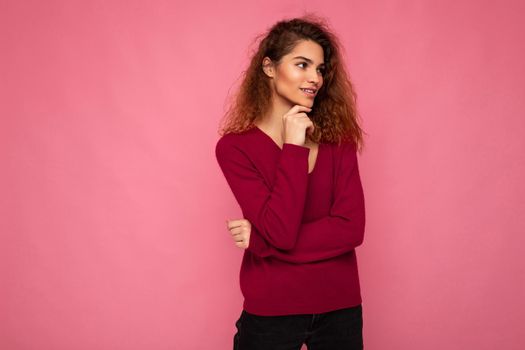 Photo of young european thoughtful fascinating brunette curly woman with sincere emotions wearing trendy pink sweater isolated over pink background with free space