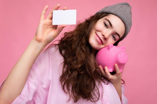 Portrait of beautiful positive cheerful cute smiling young brunette woman in stylish shirt isolated on pink background with copy space and holding pink pig moneybox and credit card for mockup