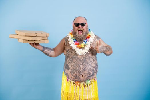 Joyful obese man with flowers garland holds boxes of pizza on light blue background