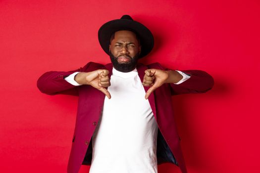 Disappointed Black man leave negative feedback, showing thumb down and grimacing from dislike and disgust, standing in party clothes over red background
