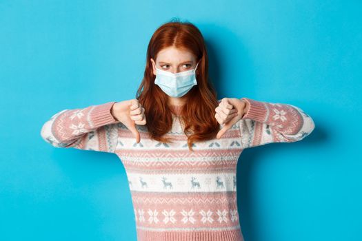Winter, covid-19 and pandemic concept. Moody redhead girl in face mask showing thumbs-down, disapprove and dislike, standing against blue background