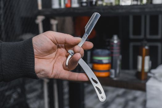 The hand of a barber or professional hairdresser holds a straight tool sharp razor equipment for shaving and beard care, close-up