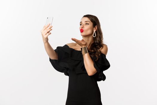 Stylish young woman in black dress, partying and taking selfie on mobile phone, sending air kiss at smartphone camera, standing over white background