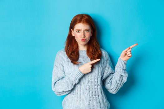 Winter holidays and people concept. Complicated redhead girl staring indecisive, pointing fingers right at logo and frowning, standing perplexed against blue background
