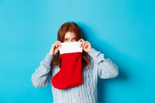 Winter holidays and gifts concept. Funny redhead girl looking inside Christmas stocking and smiling with eyes, standing against blue background
