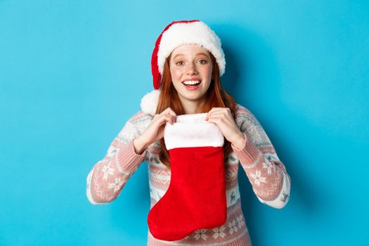 Beautiful redhead girl in santa hat open Christmas stocking and looking surprised, receiving xmas gift, standing over blue background