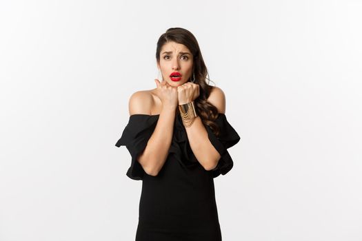 Fashion and beauty concept. Scared timid woman grimacing, looking concerned and worried at camera, standing over white background in black dress