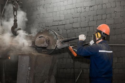 A worker in a protective helmet, respirator and overalls grinds with heavy equipment cast iron concrete tubing in the workshop of an industrial plant
