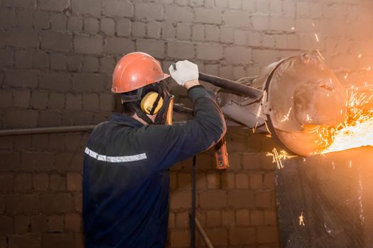 A male worker in a protective helmet, respirator, overalls manages heavy grinding equipment for cast iron concrete tubing with flying sparks in the workshop of an industrial plant