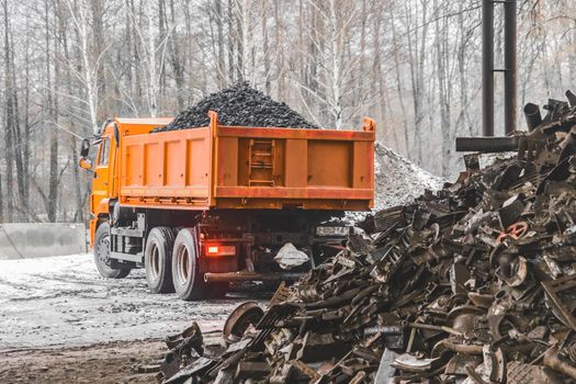 A dump truck in an industrial area or on a construction site in winter unloads coking coal from the body, next to a pile of metal waste and garbage