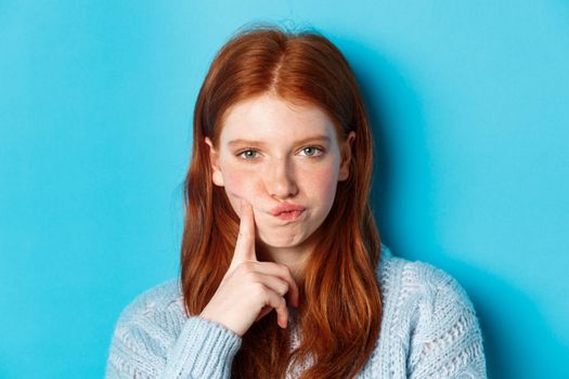 Puzzled redhead girl looking suspicious at camera, thinking or solving problem, standing against blue background