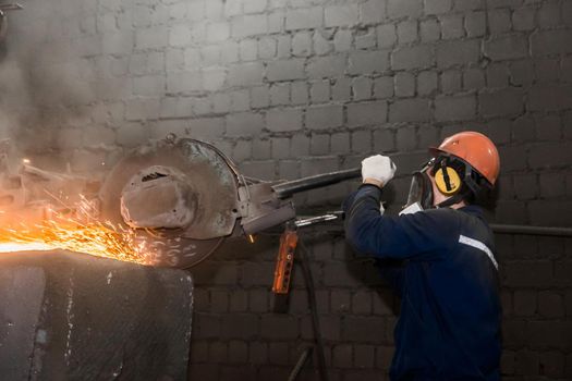A male worker in a protective helmet, respirator, overalls manages heavy grinding equipment for cast iron concrete tubing with flying sparks in the workshop of an industrial plant