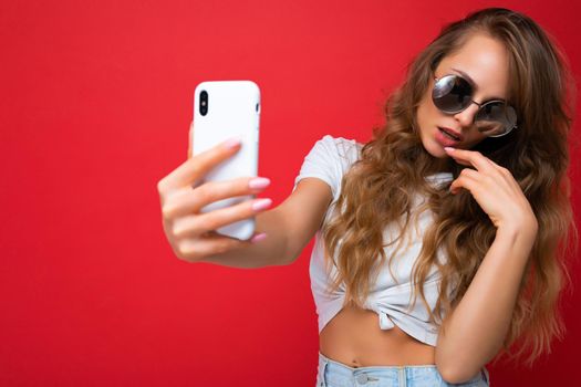 Closeup photo of amazing beautiful young blonde woman holding mobile phone taking selfie photo using smartphone camera wearing sunglasses everyday stylish outfit isolated over colorful wall background looking at device screen