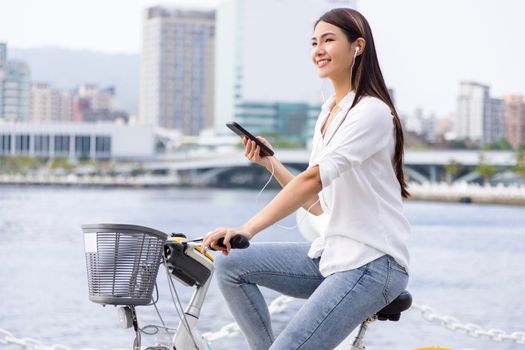 Smiling girl in headphones, holding smartphone and riding bicycle in the park