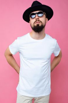 Vertical photo shot of handsome self-confident brunette unshaven young man with beard wearing casual white t-shirt for mockup black hat and stylish sunglasses isolated over pink background wall looking at camera