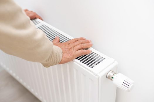 An elderly man froze in his hands, a man warms his hands near a heating radiator in an apartment, a man freezes in an apartment.