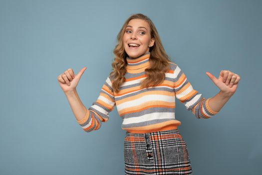 Young charming fascinating sexy positive happy smiling blonde woman wearing casual striped pullover isolated over blue background with free space and points fingers at herself