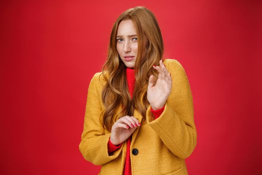 Phew disgusting take it away. Unsatisfied intense and displeased cute redhead woman in yellow coat raising hands near chest in defense stooping and grimacing from aversion over red background