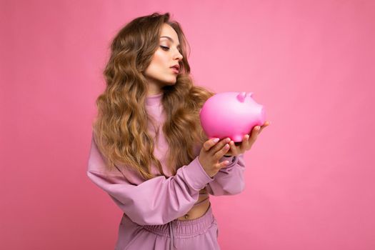 Portrait of doubtful thinking young beautiful attractive woman with blonde hair with sincere emotions wearing stylish pink hoodie isolated over pink background with copy space and holding pink piggy box. Moneybox concept