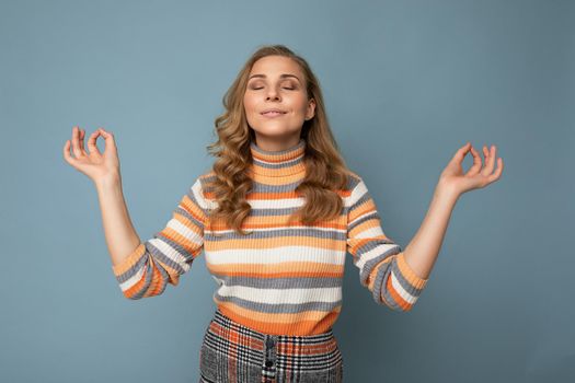 Portrait of young attractive blonde curly woman with sincere emotions wearing trendy striped pullover isolated on blue background with copy space and doing mediation gesture with fingers. Yoga concept