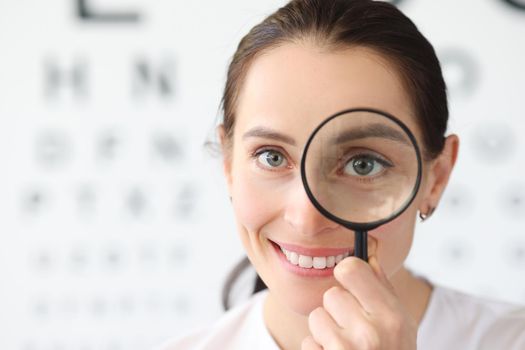 Woman optometrist holding magnifying glass in eyes against background of vision examination table
