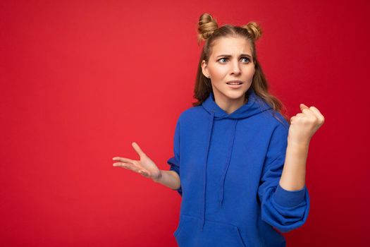 Photo of emotional agressive young beautiful blonde woman with two horns with sincere emotions wearing hipster bright blue hoodie isolated over red background with free space and showing fist