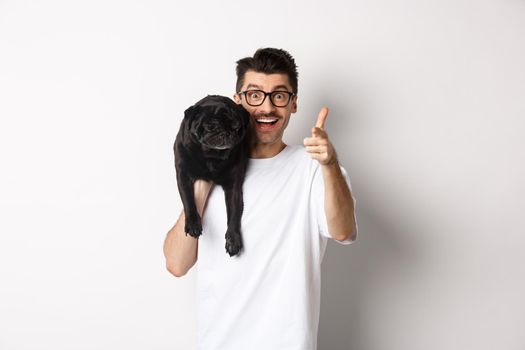 Happy young man holding cute black dog on shoulder and pointing at camera. Hipster guy carry pug on shoulder and staring at camera excited, standing over white background