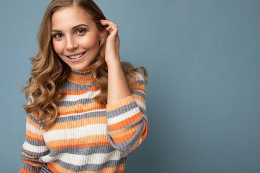 Young charming fascinating sexy positive happy smiling blonde woman wearing casual striped pullover isolated over blue background with free space