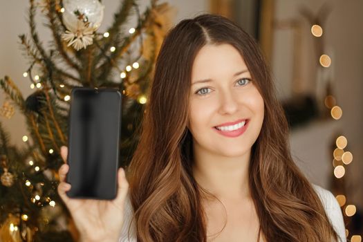 Christmas phone call and holiday greeting concept. Happy smiling woman showing screen of mobile smartphone, xmas decoration on background