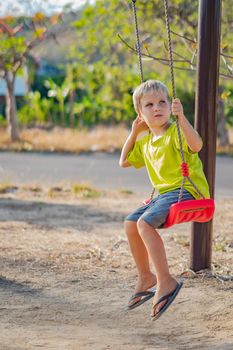 Sad lonely boy sit on swing look far away, wait for friends or while parents are busy, thinking about important things for him. Summer, childhood, leisure, friendship, relationship and people concept