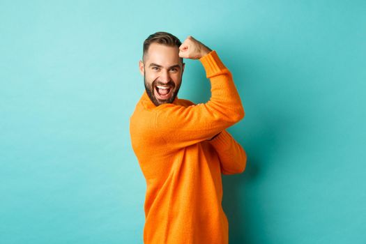 Confident handsome man show-off his muscles, flex biceps, feeling strong, standing over light blue background