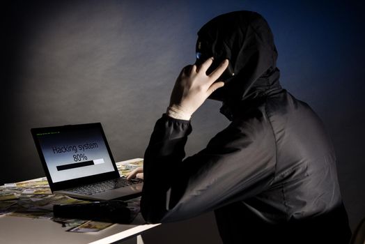 Anonymous hacker in mask sitting sideways uses a laptop and talking on the phone to hack the system in the dark. The concept of cybercrime and hacking database
