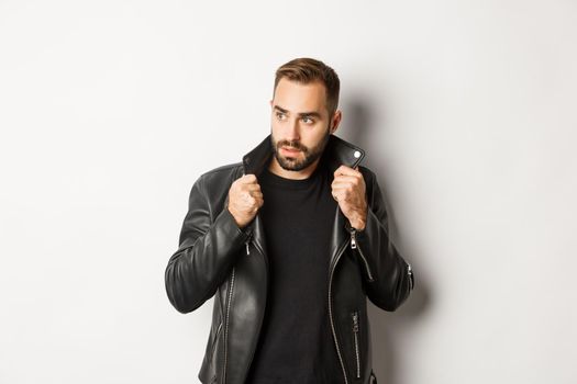 Attractive bearded man in leather biker jacket looking aside, standing confident against white background