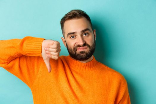 Close-up of disappointed caucasian man with beard, showing thumb down and grimacing displesed, negative feedback, standing over light blue background