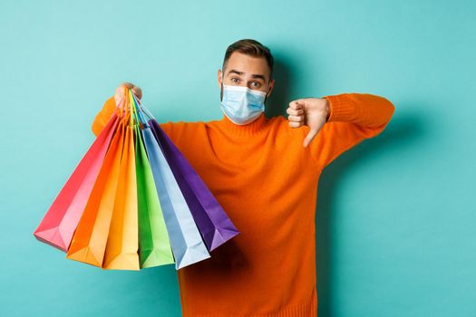 Covid-19, social distancing and lifestyle concept. Disappointed young man in medical mask from coronavirus, showing thumb down and shopping bags
