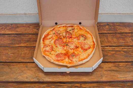 Open cardboard box packaging for freshly made pizza with cheese and salami on a wooden table background