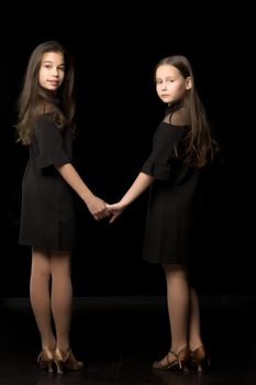 Two little girls hold hands, on a black background.