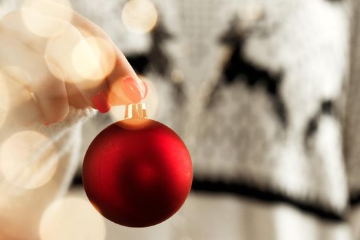 Hand of a woman in festive sweater holding Christmas bauble with blurred lights