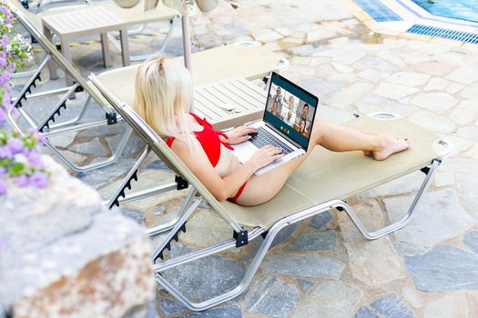 Woman Using Laptop Whilst Relaxing On Sun Lounger By Pool